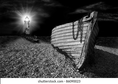 An Abandoned Wooden Fishing Boat On A Shingle Beach Illuminated At Night By The Beam From A Lighthouse In Black And White