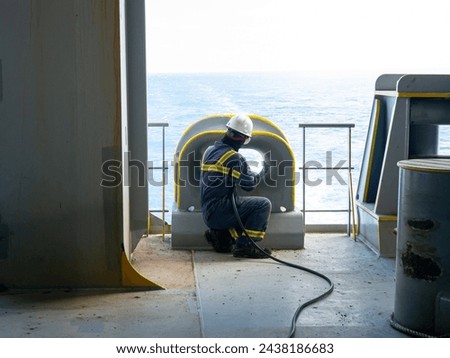 Able seaman crew member of cargo vessel is chipping rust from the ship bollard construction by air hammer and doing maintenance dressed in personal protective equipment