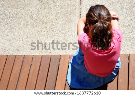 Above aerial view of sad lonely young girl (Female age 9 -10) covering her face and crying sitting on wooden floor alone in school yard. Child bullying concept. Real people. Copy space