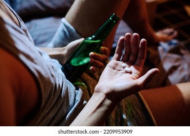 Above Angle Of Hand Of Young Depressed Man Holding Sleeping Pills Or Antidepressants And Bottle Of Alcohol While Sitting On Bed