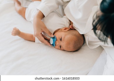 From Above Of Caring Mom Giving Pacifier To Little Asian Newborn Putting Soother Into Calm Newborn Baby Mouth Lying On Bed In House