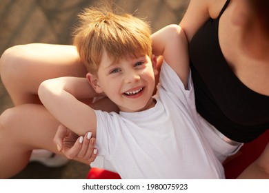 From Above Cheerful Boy Holding Hands Behind Head And Looking At Camera With Smile While Resting On Lap Of Crop Woman In Park