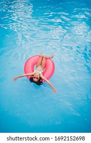 From above of cheerful little girl in swimwear and sunglasses floating on pink inflatable ring in swimming pool with blue water during summer holidays
