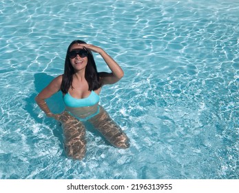 From Above Cheerful Young Hispanic Woman In Swimwear And Sunglasses Touching Forehead And Holding Hand On Hip While Kneeling In Clean Water Of Pool On Resort