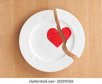 Above Closeup View Of A Broken Ceramic China White Plate With A Red Heart Painted On It.  Wood Board Background.   Concept For Relationship, Friendship Or Marriage Troubles.