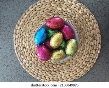 From Above Colourful Foil Wrapped Chocolate Easter Eggs In A Bowl With Straw Wicker Woven Placemat On A Polished Concrete Bench Top