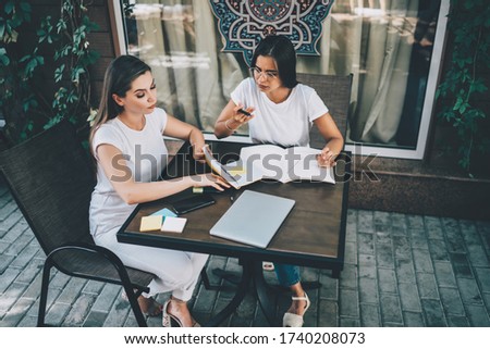 From above of concentrated female freelancers in white outfits sitting at table while working with documents and devises at cafe terrace  