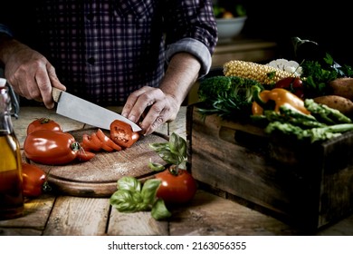 From above of crop anonymous male chef in checkered shirt slicing fresh ripe tomatoes on round shaped wooden cutting board at table with box of assorted raw vegetables in kitchen - Powered by Shutterstock