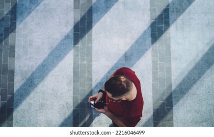 From Above Of Crop Athletic Man In Red T Shirt Browsing Smartphone On Pavement During Break And Listening To Music Through Earphones