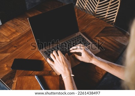 From above of crop female freelancer typing on laptop with black screen while sitting at wooden table with smartphone and notebook