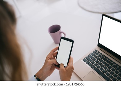 From Above Crop Unrecognizable Student Holding Smartphone With Blank Screen And Using Laptop While Sitting At White Table With Cup Of Beverage