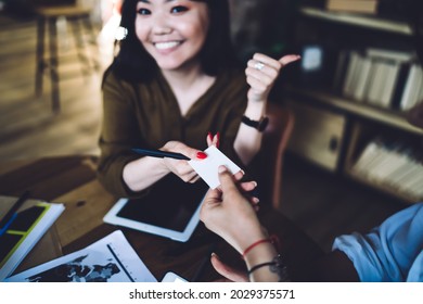 From above of crop young ethnic female employee in casual clothes with thumb up giving business card to woman and smiling at camera