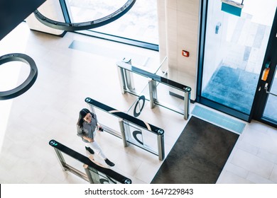 From above of dark haired female in casual shirt and white trousers passing trough turnstile in spacious bright corridor of modern building