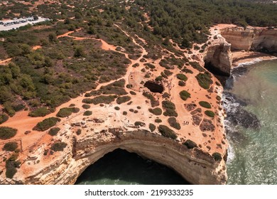 From Above Drone View Of Rough Cliff With Algar De Benagil Cave And Shrubs Located Near Splashing Water Of Clean Sea On Sunny Day In Algarve, Portugal