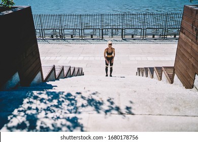 From Above Of Fit Female Athlete In Black Gym Suit Standing In Front Of Rocked Stairs And Preparing To Running Upstairs With Empty Quay On Background On Hot Summer Day 