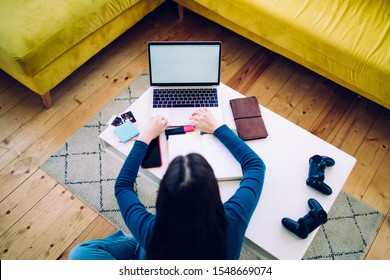 From Above Freelance Woman Sitting At Table On Floor In Living Room With Notebook And Joysticks On Top And Browsing Laptop Working At Home 