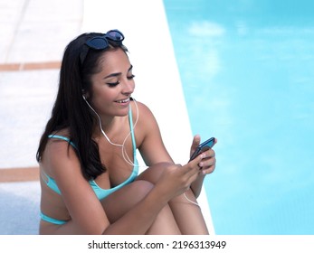 From Above Glad Young Hispanic Woman In Bikini Listening To Music And Browsing Social Media On Cellphone While Sitting Near Pool On Summer Weekend Day