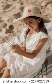 From Above Happy Child In White Dress And Straw Hat Smiling And Playing Game On Cellphone, While Sitting On Blanket On Weekend Day On Sandy Beach