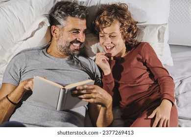 From above of happy father and son in pajamas having fun while reading interesting book and lying on soft bed together at home
