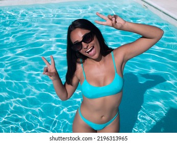 From Above Happy Hispanic Woman In Bikini And Sunglasses Smiling And Gesturing V Sign With Both Hands While Standing In Clean Pool Water On Resort