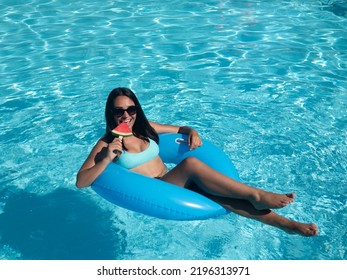 From Above Happy Hispanic Woman In Swimwear Smiling And Biting Fresh Watermelon On Stick While Floating On Inflatable Tube In Pool On Summer Weekend Day