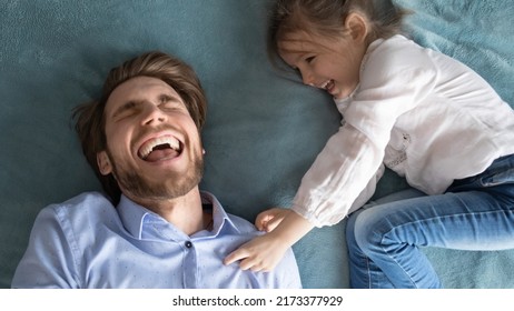Above head shot top view happy little baby girl tickling laughing young father, lying together on bed at home. Overjoyed daddy having fun with playful small kid daughter, enjoying lazy weekend pastime