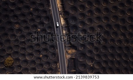 Above landscape view of black sand gound for volcanic vineyard and long straight road with white car traveling and driving on it. Concept of alternative scenic landscape outdoors