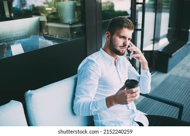 From Above Of Male Entrepreneur In Formal Clothes Looking Away While Sitting On Couch With Takeaway Coffee And Having Conversation On Smartphone