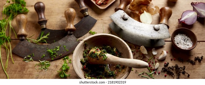 From Above Of Mortar And Pestle With Grinded Herbs Placed On Wooden Table With Old Fashioned Mezzaluna Knives In Kitchen