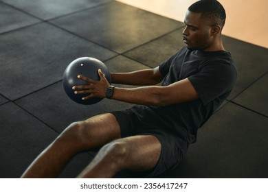 From above of motivated young African American male athlete with dark hair in black sportswear, doing Russian twist exercise with medicine ball during fitness workout in club - Powered by Shutterstock