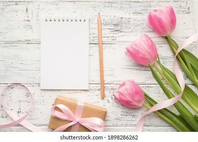 Above overhead flat lay photo of pastel color tulips on green stems wrapped giftbox with ribbon and copybook with paper blank page for mock up and pencil on wooden table