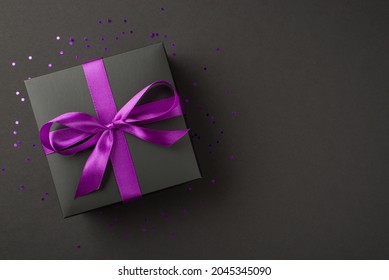 Above photo of black giftbox with purple ribbon wrapped as bow confetti and glitter around isolated on the black background