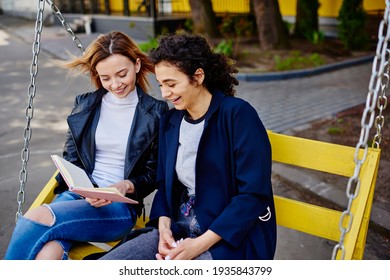 From Above Of Positive Multiracial Young Female Friends Reading Notes In Notebook And Laughing While Sitting On Yellow Wooden Swing Set In Park