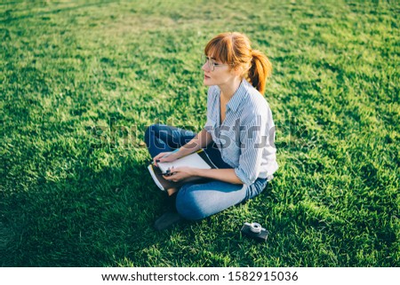 From above of red haired pensive college girl in casual clothing and eyeglasses sitting with crossed legs on green grass while learning lessons with notebooks at park 