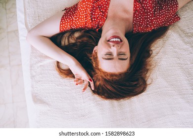 From above satisfied happy red-haired woman in red dotted dress relaxing on soft cozy bed while daydreaming with closed eyes - Shutterstock ID 1582898623