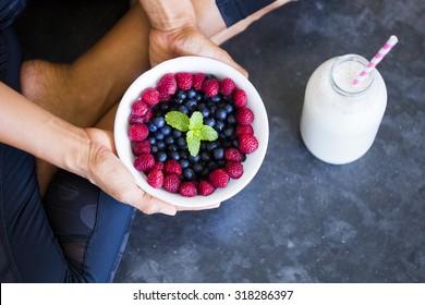 Above shot of the hands of a woman with a bowl of berries and a bottle of almond milk wearing a sportive outfit.