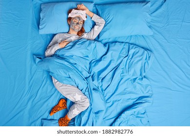 Above Shot Of Happy Redhead Young European Woman Dressed In Casual Pajama Feels Comfortable Applies Clay Mask On Face For Reducing Wrinkles Wakes Up Early In Morning. People And Bed Time Concept