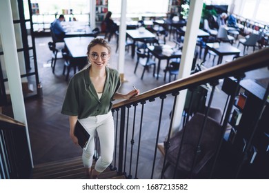 From Above Smart Student Woman In Glasses Carrying Book While Climbing Up Stairs Holding Railing And Smiling At Camera In Light Library
