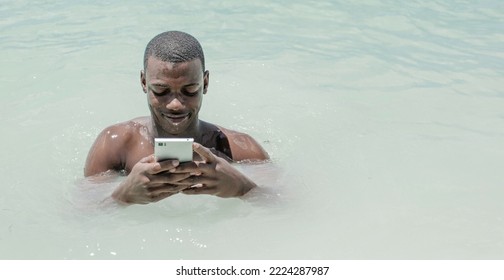 From Above Smiling Black Male Traveler Sitting Inside The Sea And Browsing Social Media On Cell Phone During Summer Vacation At Resort.