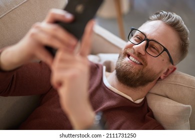 From Above Of Smiling Male Hipster Lying On Couch And Messaging On Mobile Phone While Relaxing At Weekend At Home