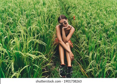 From Above Of Stylish Young Ethnic Millennial In Sneakers And Sunglasses Sitting In Tall Grass And Recreating During Summer Holidays In Countryside