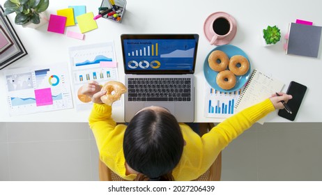 Above top view of asia girl or woman staff worker sitting busy work in multi task job or relax study MBA at home office on desk eat bite diet cake fast food late lunch meal in hurry break coffee time.