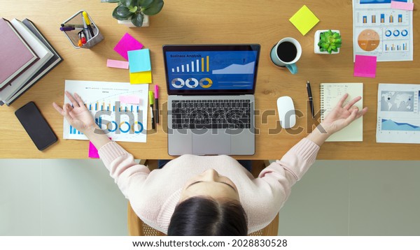 Above top view of asia girl work on desk table\
home office sit calm inhale deep stress relief in zen lotus yoga\
post. Good break rest time for teen labor people serene life in job\
career workforce.