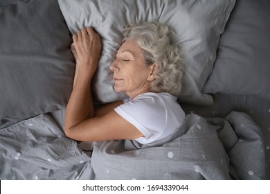 Above Top View Calm Peaceful Elderly Mature Hoary Woman Sleeping On Soft Pillow Under Blanket, Enjoying Sweet Dreams At Night. Happy Middle Aged Granny Lying On Side, Resting In Comfortable Bed Alone.