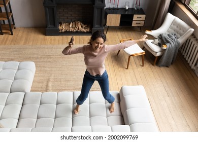 Above Top View Emotional Energetic Young Hispanic Woman Jumping Barefoot On Huge Couch, Listening Disco Music In Mobile Application, Having Fun Entertaining Alone In Living Room, Celebrating Success.