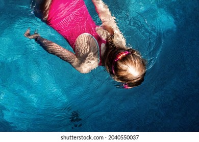 Above Top View Of Little Cute Kid Girl In Sport Pink Swimsuit And Goggles Diving Underwater In Clear Blue Water Of Indoor Swimming Pool At Dark Evening Time. Children Watersport Healthy Activities