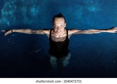 Above top view portrait of young adult caucasian female model in sport black swimsuit stand in clear blue water of indoor swimming pool at dark evening time. Relax harmony and wellness spa therapy