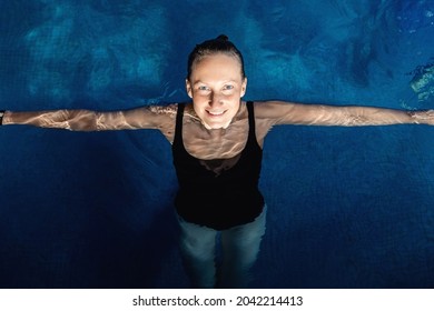 Above Top View Portrait Of Young Adult Caucasian Female Model In Sport Black Swimsuit Stand In Clear Blue Water Of Indoor Swimming Pool At Dark Evening Time. Relax Harmony And Wellness Spa Therapy