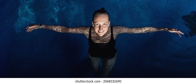 Above Top View Portrait Of Young Adult Caucasian Female Model In Sport Black Swimsuit Stand In Clear Blue Water Of Indoor Swimming Pool At Dark Evening Time. Relax Harmony And Wellness Spa Therapy