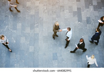 From Above Two Adult Business Men Shaking Hands Met In Office Hall With Workers Walking Around.